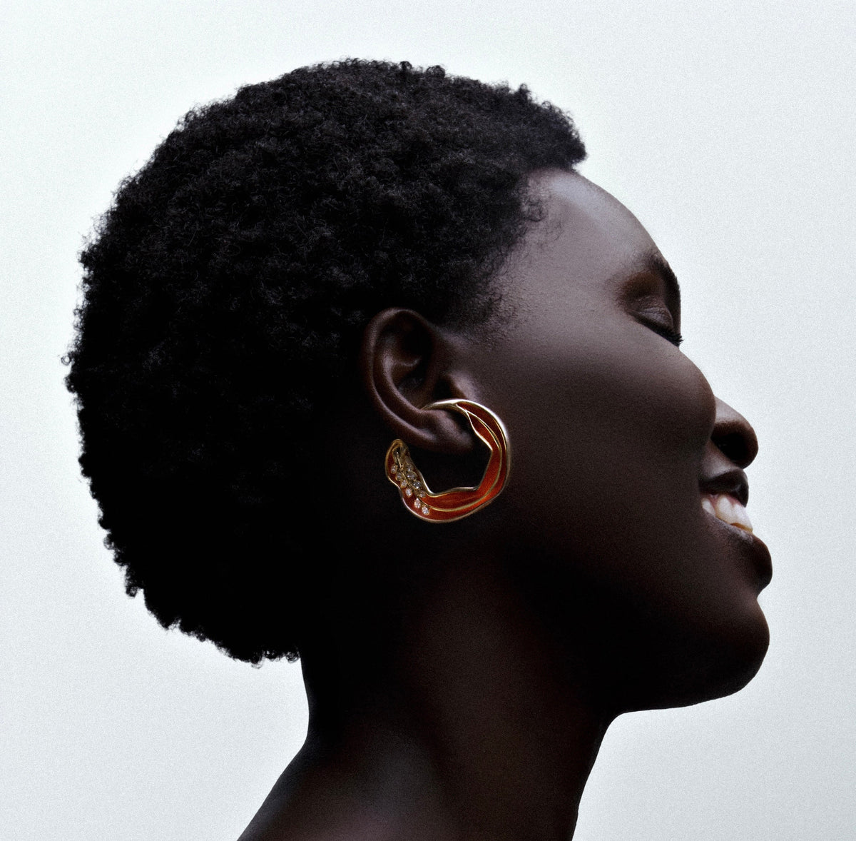 A close-up side profile of a person with short dark curly hair wearing a large gold hoop earring with small clear stones, against a light gray background.