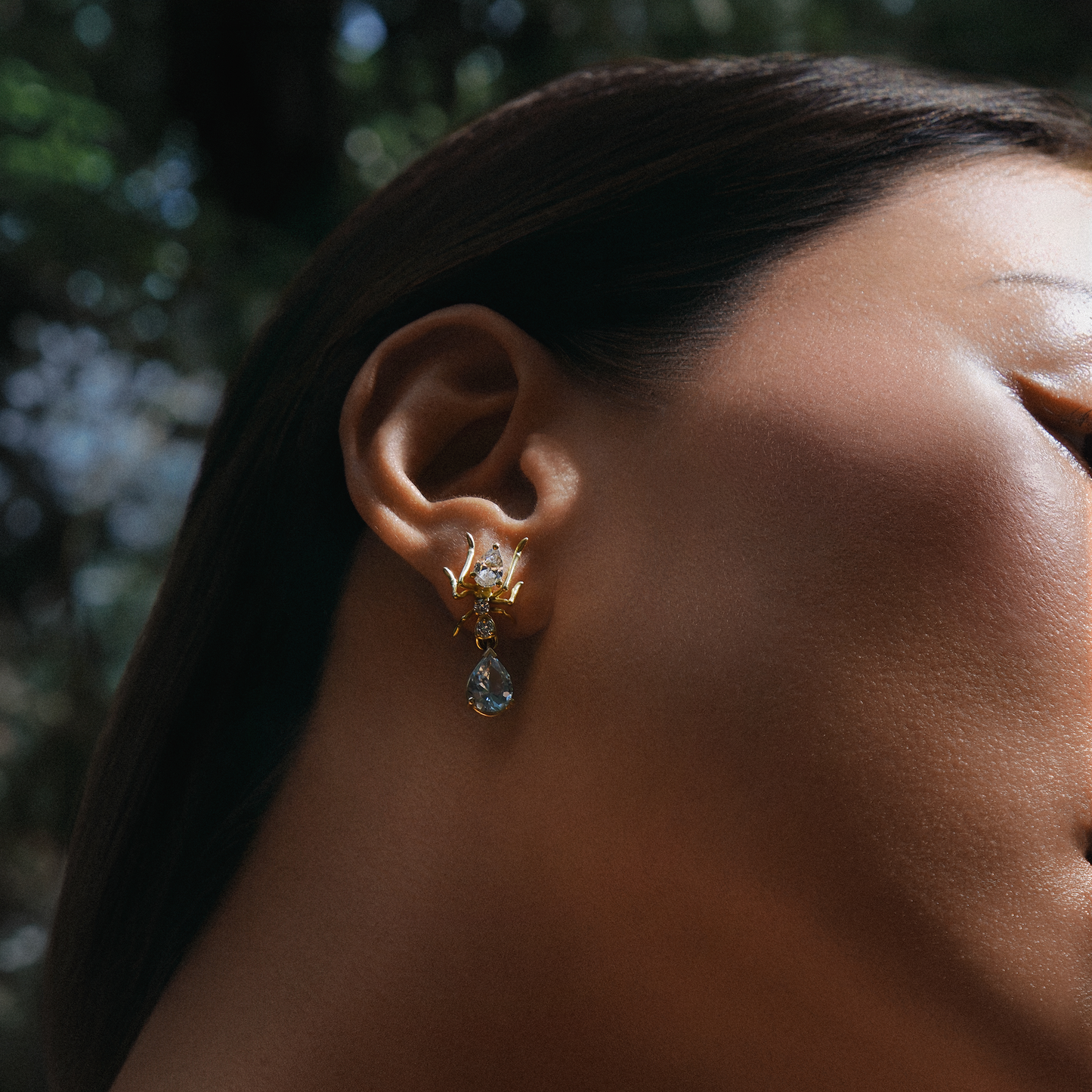 A close-up side profile of a woman wearing a delicate gold huggie hoop earring with a small gold leaf cluster and a teardrop-shaped light blue gemstone dangle, set against a blurred outdoor background.
