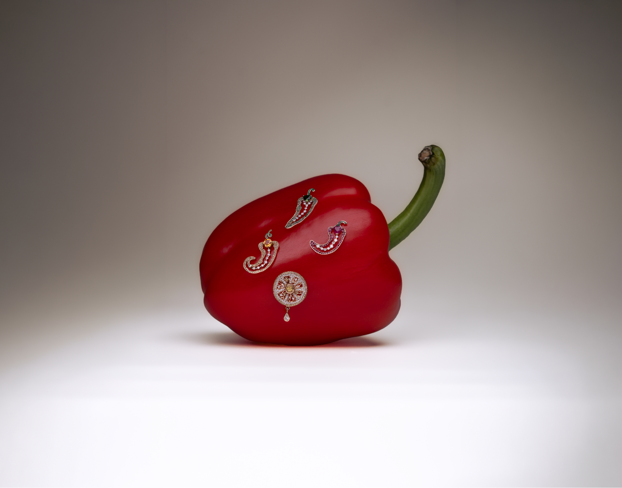 A glossy red bell pepper-shaped box with a green stem, decorated with small metallic and gemstone embellishments, set against a neutral gray studio background.